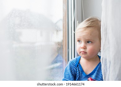 Little Sad Boy Sitting At Home On Window In Rainy Day. Stay Home Coronavirus Concept.