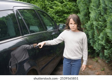 A little sad girl in glasses, a sweater and jeans stands near a family car. Insurance and safety concept - Powered by Shutterstock