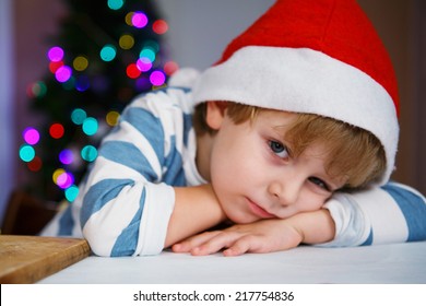 Little Sad Or Serious Child In Santa Hat With Christmas Tree And Lights On Background