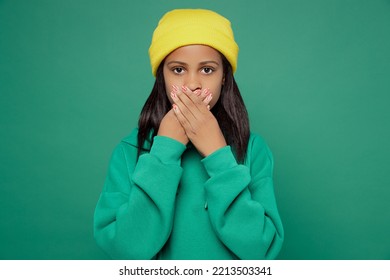 Little Sad Shocked Scared Kid Teen Girl Of African American Ethnicity 13-14 Years Old Wear Casual Hoody Hat Cover Mouth With Hand Look Camera Isolated On Plain Dark Green Background Childhood Concept