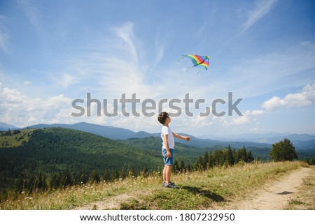 Little school age boy running down the slope with kite in the sun. Sunny summer or spring day at sunset. Active outdoor games and leisure. Mountain landscape with hills and coniferous trees.