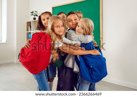 Little school best friends are happy to meet in school classroom after school holidays. Happy preteen boys and girls with backpacks hugging while standing near blackboard and looking at camera.