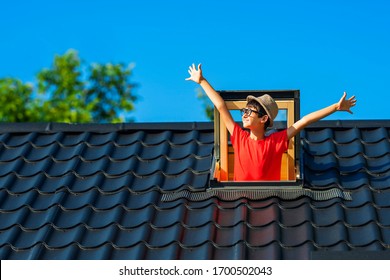 Little school boy celebrates the beginning of summer vacation, new day, morning. Happy childhood, sunny summer time. Roof is covered with metal tiles. Child looks out the window. Blue sky background.