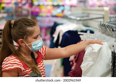   Little School Girl In A Protective Medical Mask Chooses School Uniform In A Store.  Preparing For School. Prevention Of Coronavirus. Back To School Shopping.                                