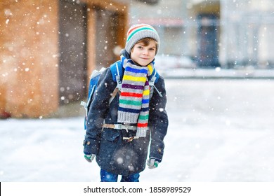Kleiner Schüler der Grundschule, der während des Schneefalls zur Schule geht. Fröhliches Kind, das Spaß hat und mit dem ersten Schnee spielt. Student mit Rucksack oder Satchel in bunten Winterkleidung.