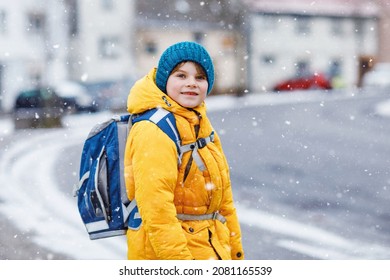 Little school kid boy of elementary class walking to school during snowfall. Happy child having fun and playing with first snow. Student with in yellow jacket and backpack in colorful winter clothes.