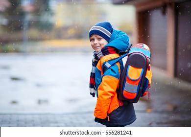 Little School Kid Boy Of Elementary Class Walking To School During Snowfall. Happy Child Having Fun And Playing With First Snow. Student With Backpack In Colorful Winter Clothes Catching Snowflakes.