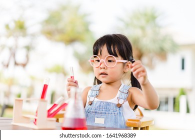 Little Scientist Asian Toddler Nerd Girl Wearing Glasses With Red Liquid Into Flask And Shaking.Home School Girl Learning About Science And Pouring Reagent Into Flask.Chemistry Class At Home.Nerd Kid.