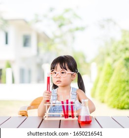 Little Scientist Asian Toddler Nerd Girl Wearing Glasses With Red Liquid Into Flask And Shaking.Home School Girl Learning About Science And Pouring Reagent Into Flask.Chemistry Class At Home.Nerd Kid.