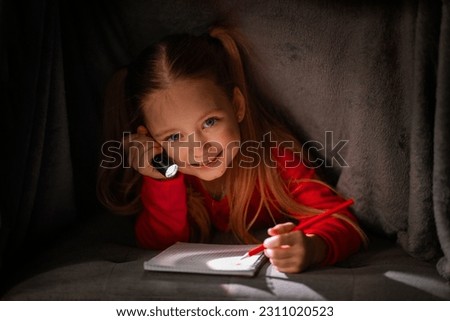 Little Secrets. Cute Smiling Girl Hiding Under Blanket With Flashlight And Writing In Notebook, Happy Preteen Female Kid Relaxing Under Covers, Holding Pocket Torch and Taking Notes To Notepad