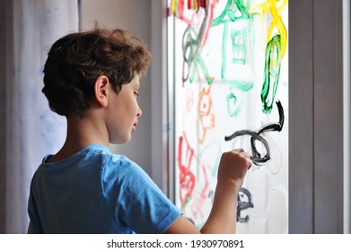 A little serious boy holds a brush in his hands and draws pictures on the window in his room. The child draws a rainbow, a house and an abstraction.