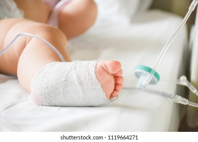 Little Sick Baby On A Drip Receiving A Saline Solution Intravenously In The Hospital Or Clinic. Baby With Cannula In The Feet On A Hospital Bed