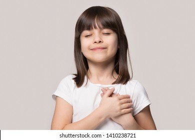 Little Sincere Adorable Girl Closed Eyes Holding Hands On Chest Feeling Gratitude Pose Isolated On Sandy Color Beige Background, Arms On Heart Gesture Of Love Appreciation Gratitude, Adoption Concept