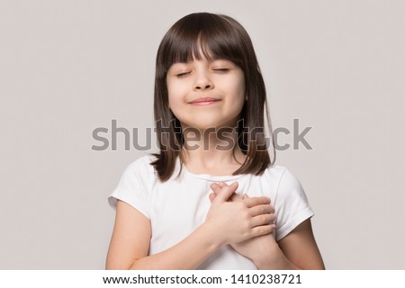 Little sincere adorable girl closed eyes holding hands on chest feeling gratitude pose isolated on sandy color beige background, arms on heart gesture of love appreciation gratitude, adoption concept