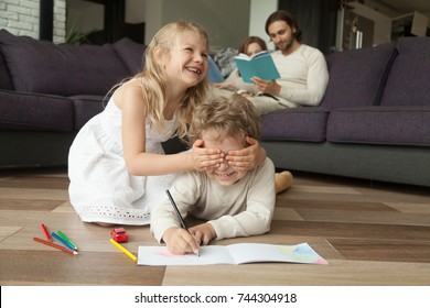 Little sister closing brothers eyes with hands, siblings playing peek a boo laughing on floor, parents reading book on sofa at home, family with children spending leisure time together in living room