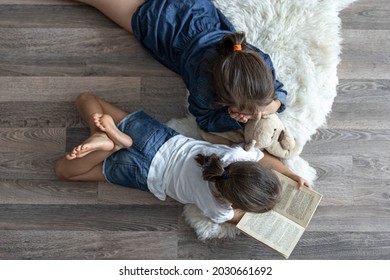 Little Sisters Are Reading A Book With A Teddy Bear Lying On The Floor In The Room Top View.