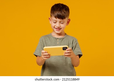 Little Small Boy 6-7 Years Old In Green T-shirt Using Play Racing App On Mobile Cell Phone Gadget Smartphone For Pc Video Games Isolated On Plain Yellow Background. Mother's Day Love Family Concept