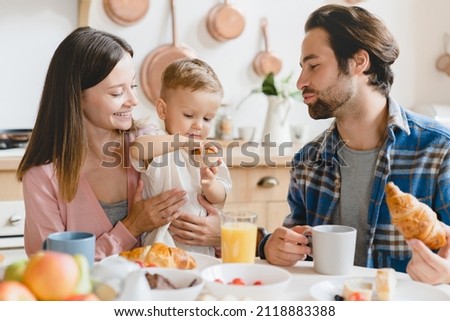 Little small cute son daughter child kid toddler new born infant eating cake during breakfast with young parents. Mother and father feeding child at home kitchen