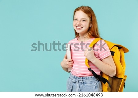 Little smart happy pupil redhead kid girl 12-13 year old in pink striped t-shirt hold notebook book yellow school bag backpack isolated on pastel blue background Children lifestyle childhood concept