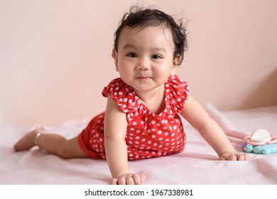 Little Smiling Asian Baby, A Cute Toddler Dressed In Red Practicing Crawling On The Bed And Looking At The Camera