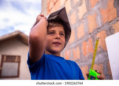 A Little Smiling Boy With A Newspaper Hat Holds A Measuring Tape In His Hands