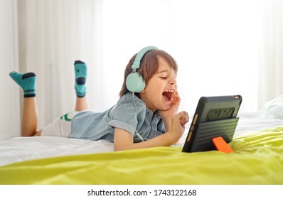 Little smiling boy siting on bed and playing on a digital tablet at home. Kid in his bedroom wearing headphones and using smart devices having fun online with his friends in a video call - Powered by Shutterstock