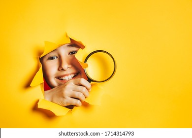 Little smiling caucasian boy looks through torn hole in paper yellow wall, holding magnifying glass in his hand. Search, discovery, research concept. Sidebar for text information, advertising, event.