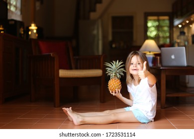 Little Smiling Female Kid Sitting On Floor In Living Room With Pineapple And Showing Thumbs Up, Laptop In Background. Concept Of Health Life, Fruit And Childhood.