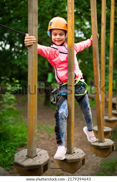 Little smiling girl in helmet and harness\
passing on trail in sky rope park in\
summer