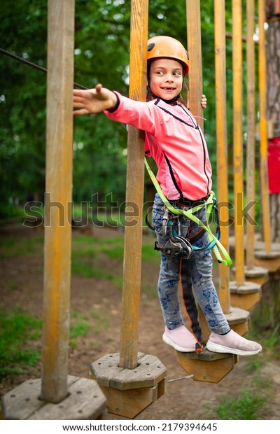 Little smiling girl in helmet and harness\
passing on trail in sky rope park in\
summer