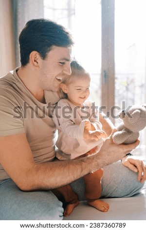 Little smiling girl sits on the lap of a laughing dad with a teddy bear in his hand