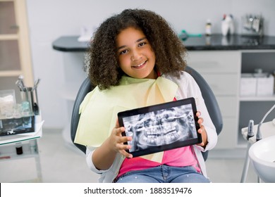 Little smiling mixed raced girl with curly hair sitting in dental chair and looking at camera, while holding x-ray scan image of her teeth on digital tablet. Pediatric dentistry, orthodontics