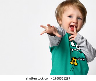 Little Smiling Preschool Toddler Boy European Ethnicity Wearing Trendy Green Raglan With Cute Monster Muzzle Print And Sportive Black Pants Stand With Hand Reaching To Camera. Studio Portrait On White