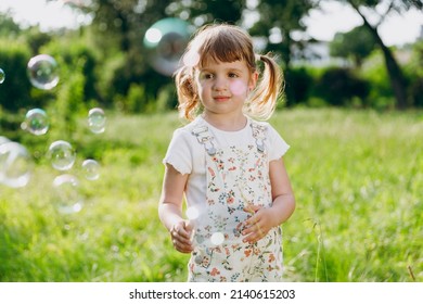 Little smiling satisfied happy kid girl 5-6 years old in white casual clothes blowing bubbles play on park green sunshine lawn, spending time outdoor in village countyside during summer time vacations - Shutterstock ID 2140615203