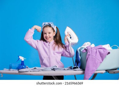 Little Smiling Sweet Girl Irons Clothes With Iron, Strongwoman Raises Hand To Show Biceps, Child Defeated A Mountain Of Laundry Items.