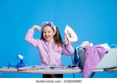 Little Smiling Sweet Girl Irons Clothes With Iron, Strongwoman Raises Hand To Show Biceps, Child Defeated A Mountain Of Laundry Items.