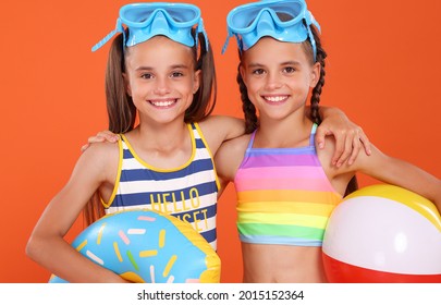 Little Smiling Twin Sisters Wearing Diving Goggles, Embracing Each Other, Holding Inflatable Beach Ball And Donut, Ready For Family Vacation At Seaside, Isolated On Orange Studio Background