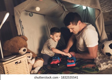 Little son plays in cars with dad in a toy house. Cheerful dad lies with his son in the house and plays toys at night. 