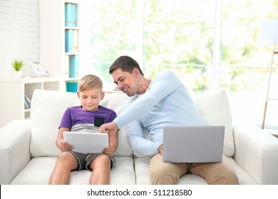 Little son with tablet and father with laptop sitting on sofa at home