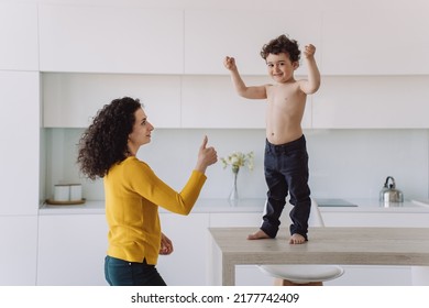 Little Spanish Curly Boy In Blue Pants Standing On A Table Showing His Biceps, Smiling, Her Mom Looking At Him With Proud Gesturing Thumb Up At Kitchen. Healthy Kid Rising Fists Up In A Winner Gesture