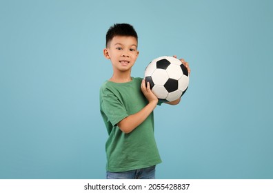 Little Sportsman. Excited Asian Boy Posing With Football Ball In Hands Over Blue Background In Studio, Cute Preteen Korean Male Kid Looking At Camera, Enjoying Playing Soccer, Copy Space