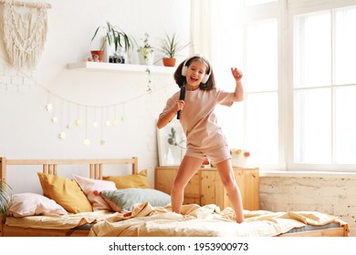 Little star. Cute happy little girl wearing pajama with hairbrush as mic pretending to be famous singer while standing on bed at home, adorable preschool child singing and dancing in bedroom