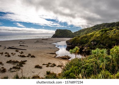 A little stream runs to the ocean on a remote beach on a cloudy day with rocks and cliffs on the background. West Coast, South Island, New Zealand.
