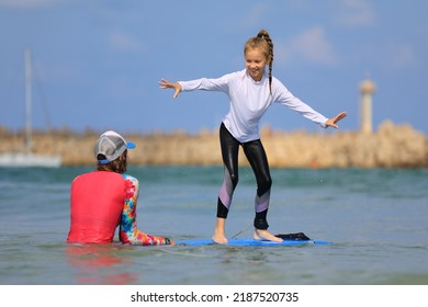 Little Surf Girl - Young Surfer Learn To Ride On Surfboard With Instructor At Surfing School. Active Family Lifestyle, Kids Water Sport Lessons, Swimming Activity In Summer Camp. Vacation With Child.