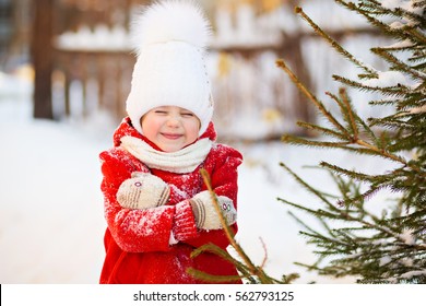a little sweet, beautiful little girl 4 to 5 years old in a red coat, white hat with a fluffy pom-pom winter walks on the street next to the fir, frozen, eyes closed smiling
