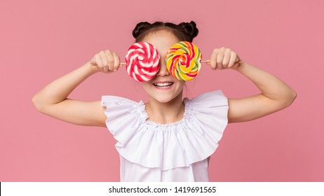 Little sweet tooth. Adorable girl covering her eyes with two colorful lollipops, pink panorama background - Powered by Shutterstock