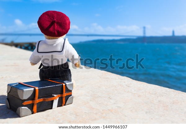 Little teddy bear wear sailor suit\
and beret hat, sitting on suitcase at quay in Lisbon, Portugal,\
view to bridge across water, dreaming of travel (copy\
space)