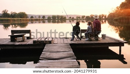 Little teen boy and girl with their grandpa sitting at the lake pier with rods and fishing equipment and talking early in the morning. Fishing and relationships concept 