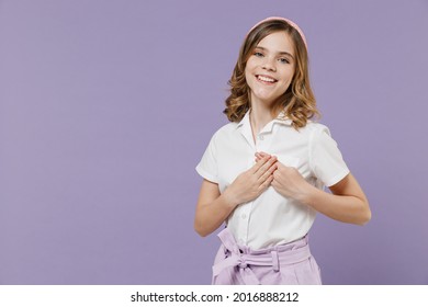 Little thankful happy kind blonde kid girl 12-13 years old in white short sleeve shirt put folded hands on heart isolated on purple background children studio portrait. Childhood lifestyle concept.