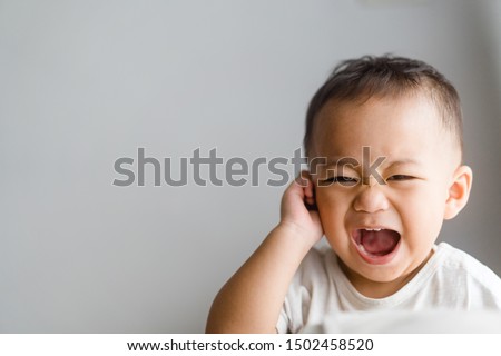 Little toddler boy has earache when insects inside isolated on grey background.2.6 years old baby boy hands touching in ear and screaming.